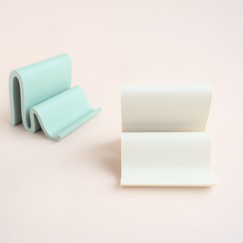May include: Two minimalist phone stands. One is a light teal color with a curved, wave-like design. The other is white with a textured surface and a similar curved shape. Both are on a light pink background.