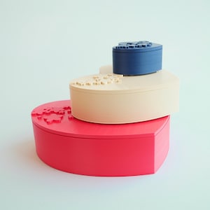 Może przedstawiać: Trzy pudełka w kształcie serca ułożone jeden na drugim. Dolne pudełko jest czerwone, środkowe beżowe, a górne niebieskie. Wieczka mają wzory puzzli.