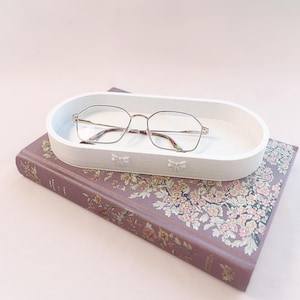 Peut inclure: Un plateau ovale blanc avec des détails de nœuds contient une paire de lunettes à monture dorée. Le plateau repose sur un livre violet avec des illustrations florales.