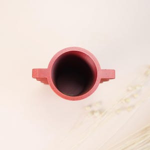 May include: A top-down view of a red cylindrical vase with two small, rectangular handles. The vase has a dark interior and is set against a light pink background. The vase is likely made of concrete or a similar material.