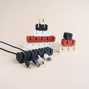 May include: Cable organizers in black, white, red, and beige. The organizers are stacked in various configurations, with USB cables inserted. The background is a solid, light beige color. These organizers help manage and organize cables.