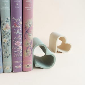 May include: A row of colorful books with floral and peacock feather designs, next to two heart-shaped bookends. One bookend is light green, the other is off-white. The books are part of the Cranford Collection.