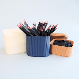May include: A selection of stationery containers in different colours. A navy blue pencil holder is filled with black pencils with pink tips. A peach-coloured container holds black items, and a cream-coloured container sits to the left.