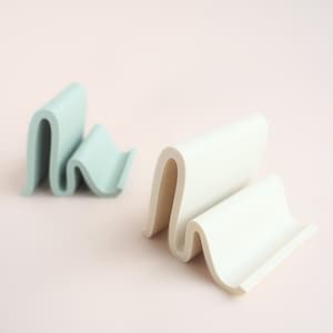 May include: Two wavy, minimalist soap dishes. One is a soft, muted green, and the other is off-white. The design features a series of curved lines, creating a modern and functional aesthetic. The soap dishes are made of a smooth material.