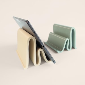 Könnte beinhalten: Ein Tablet ruht in einem wellenförmigen Ständer in Beige. Ein zweiter, ähnlicher Ständer in gedämpftem Grün ist ebenfalls sichtbar. Die Ständer sind so konzipiert, dass sie Tablets aufrecht halten. Das Tablet ist dunkelgrau.