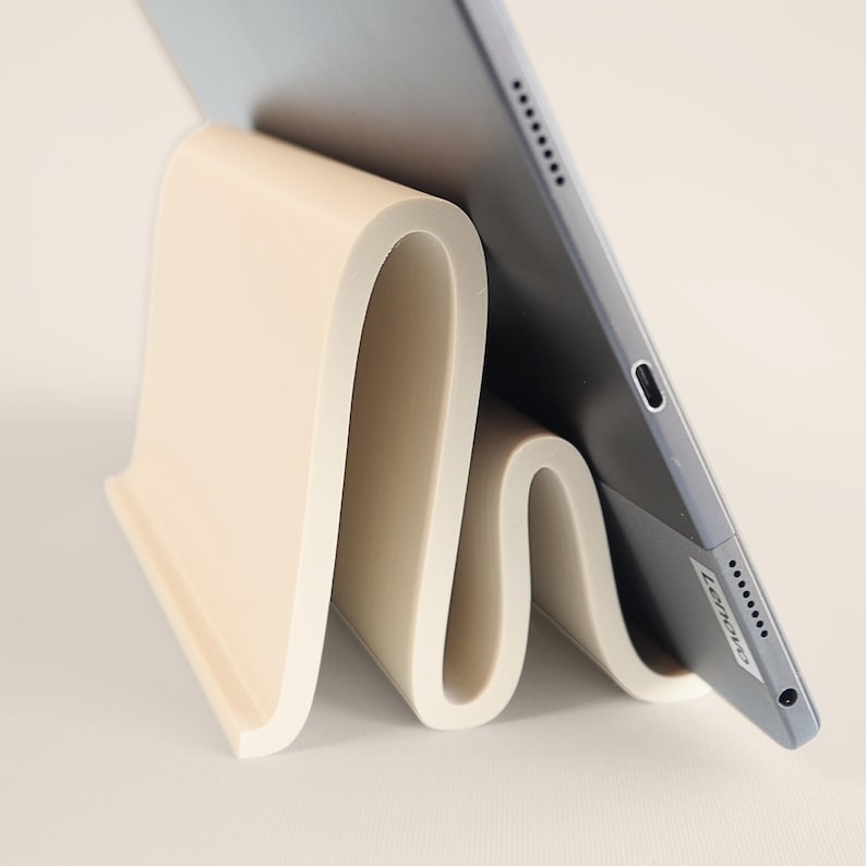 May include: A cream-colored tablet stand with a unique, wavy design. The stand is holding a gray tablet, showcasing its sleek form. The stand's minimalist aesthetic complements the tablet, offering a stylish and functional accessory for any desk or workspace.