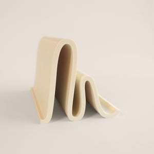 May include: A cream-colored, wavy sculpture with a smooth, matte finish. The design features a continuous, undulating form, creating a sense of movement and fluidity. The object is set against a neutral background, highlighting its unique shape.