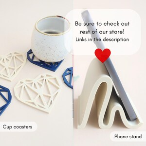 May include: The image showcases a collection of heart-shaped cup coasters in white and blue, alongside a white phone stand. A ceramic cup sits atop a blue coaster. Text reads: "Be sure to check out rest of our store! Links in the description."
