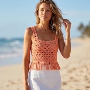 Peut inclure: Un haut sans manches en crochet de couleur pêche avec une encolure festonnée, des motifs géométriques ajourés complexes et un ourlet droit terminé par de longues franges pendantes. Le vêtement est associé à une jupe blanche unie.
