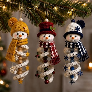 Peut inclure: Trois décorations de bonhomme de neige en crochet suspendues à une branche. Le bonhomme de neige de gauche porte un bonnet et une écharpe jaune moutarde avec des étoiles jaunes. Celui du centre a un haut-de-forme rouge et une écharpe avec du houx. Celui de droite a un bonnet et une écharpe bleu marine avec des flocons de neige bleus.