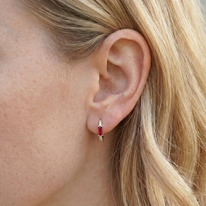 Könnte beinhalten: Nahaufnahme eines Ohres einer Frau mit einem kleinen goldenen Creolen-Ohrring. Der Ohrring hat einen rechteckigen, tiefroten Edelstein und kleine weiße Steine. Die Frau hat blondes Haar.