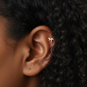 May include: A close-up of a person's ear with a small, gold-colored dragonfly earring. The earring is positioned on the upper ear cartilage. The person has dark, curly hair.