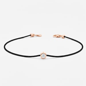 Könnte beinhalten: Ein filigranes Armband mit schwarzem Band und roségoldenen Verschlüssen. Ein einzelner, runder Diamant ist in einer roségoldenen Lünette in der Mitte gefasst. Das Armband ist minimalistisch und elegant.