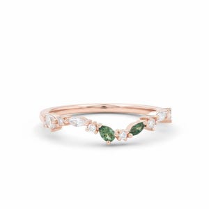 May include: Rose gold ring with a curved design. The ring features a series of white and green gemstones. The gemstones are a mix of marquise and round cuts. The ring is set against a white background.