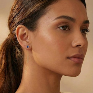 Könnte beinhalten: Nahaufnahme eines Ohrs einer Frau mit einem kleinen, runden, blauen Edelstein-Ohrstecker. Eine zarte Goldkette hängt von dem Ohrstecker herab und endet in einem dünnen, rechteckigen Stab. Die Frau hat braunes Haar, das zu einem Pferdeschwanz gebunden ist.