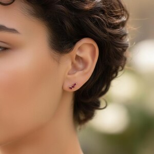 Könnte beinhalten: Nahaufnahme eines Ohrs mit einem goldenen Ohrring. Der Ohrring hat drei ovale, tiefrote Edelsteine, die in einem geschwungenen Design angeordnet sind. Das Design ist dezent und elegant.