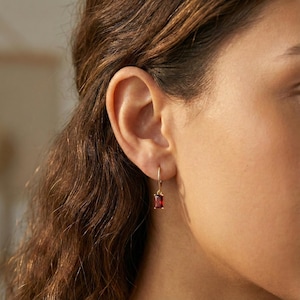 Könnte beinhalten: Nahaufnahme eines goldenen Ohrrings mit einem rechteckigen, tiefroten Edelstein. Der Ohrring ist ein einfacher Tropfenstil, wobei der Edelstein in einen goldenen Rahmen gefasst ist. Der Hintergrund ist unscharf.