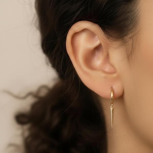 May include: Close-up of a gold hoop earring with a pointed, spike-like charm. The earring is hanging from an ear. The earring is a simple, elegant design.