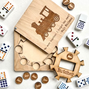 May include: Wooden game set with a train engine design and the quote "We were together. I forget the rest." - Walt Whitman. The game includes a wooden box with a train engine design and a separate wooden piece with a star shape and the word "Butcher" on it. The game also includes dominoes and small wooden train engine pieces.