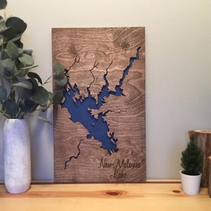 May include: Wooden wall art featuring a detailed map of New Melones Lake, with a blue lake cutout against a brown wood grain background. The text "New Melones Lake" is carved at the bottom. A compass rose is at the top.