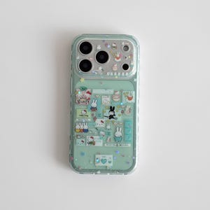 Könnte beinhalten: Eine mintgrüne Handyhülle mit einem transparenten, glitzernden Overlay. Die Hülle zeigt verschiedene Aufkleber von Zeichentrickfiguren, darunter Hasen und Hello Kitty. Die Kameralinsen sind oben sichtbar.