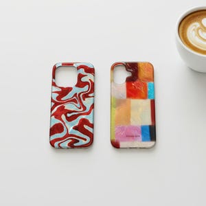 Puede incluir: Dos fundas para teléfono con diseños coloridos. Una funda tiene un patrón de remolino rojo y azul. La otra funda presenta un mosaico de varios colores, incluyendo rojo, naranja, amarillo y azul. Una taza de café blanca con arte latte en el fondo.
