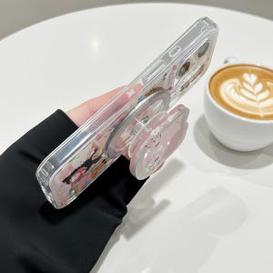 May include: Clear phone case with a pink pop-up grip. The case features a colorful design with cartoon characters. The pop-up grip is pink and has a cartoon character design. The phone case is held at an angle.