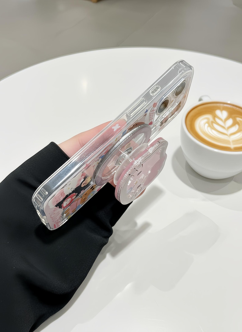 May include: Clear phone case with a pink pop-up grip. The case features a colorful design with cartoon characters. The pop-up grip is pink and has a cartoon character design. The phone case is held at an angle.