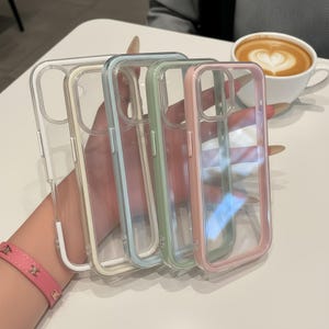 May include: Four clear phone cases with colored borders in white, cream, light blue, green, and pink. The cases are held up, showcasing their transparent design and the colored edges. The cases are designed to protect the phone.