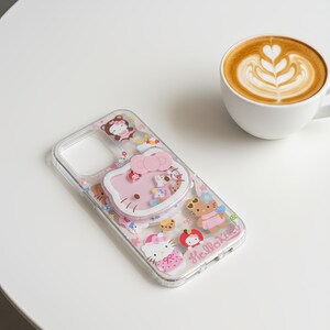 May include: Clear phone case with a pink Hello Kitty design and various character illustrations. The case features a pop-up grip and the words "Hello Kitty". A cup of coffee with latte art is in the background.
