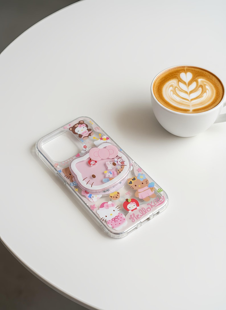 May include: Clear phone case with a pink Hello Kitty design and various character illustrations. The case features a pop-up grip and the words "Hello Kitty". A cup of coffee with latte art is in the background.