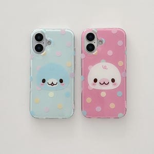 Könnte beinhalten: Zwei Handyhüllen, eine hellblaue und eine rosa, jeweils mit einem Cartoon-Robben-Charakter und bunten Punkten verziert. Die Hüllen haben ein klares, glänzendes Finish und sind für ein Smartphone konzipiert.