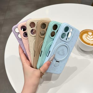 Pode incluir: Uma coleção de capas de telefone coloridas em tons pastel de lavanda, rosa, bege, verde menta e azul claro. Cada capa apresenta um suporte de anel com um design e o texto "LOVELY TIME". As capas têm um padrão de bolinhas.