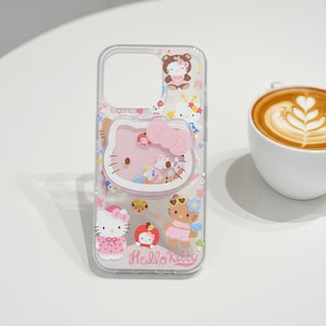 May include: Clear phone case featuring a pink Hello Kitty design with a bow and various character illustrations. The case has a transparent border and includes the text "Hello Kitty" at the bottom. The design incorporates pink, white, and other pastel colors.
