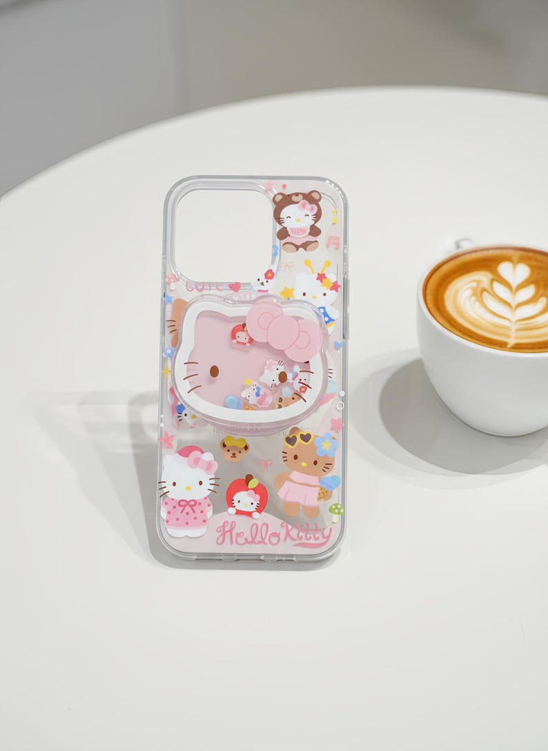May include: Clear phone case featuring a pink Hello Kitty design with a bow and various character illustrations. The case has a transparent border and includes the text "Hello Kitty" at the bottom. The design incorporates pink, white, and other pastel colors.