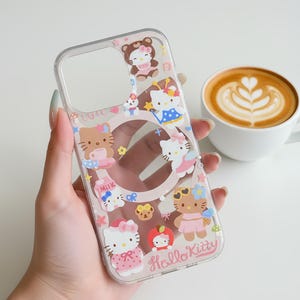 May include: Clear phone case featuring Hello Kitty and other cartoon characters in various outfits. The case has a pink border and the words "Hello Kitty" are printed on the bottom. A cup of coffee with latte art is in the background.