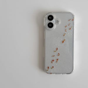 May include: Clear phone case with a glittery silver background and a pattern of small, orange and gold fish. The case has a clear border and a camera cutout.