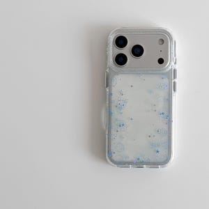 Könnte beinhalten: Transparente Handyhülle mit silbernem Kameramodul. Die Hülle hat ein Design aus blauen Cartoonfiguren und Sternen. Die Handyhülle besteht aus transparentem Material.