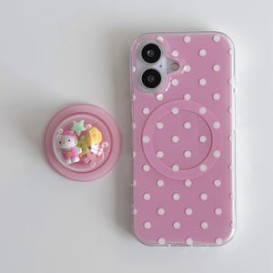 Könnte beinhalten: Rosa Handyhülle mit weißen Punkten und transparentem Rand. Ein passender rosa Handygriff mit Hello Kitty-Design befindet sich neben der Handyhülle. Die Handyhülle hat ein kreisförmiges Design auf der Rückseite.