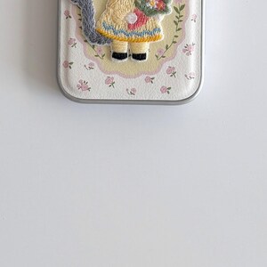 a small, decorative case with a floral pattern and a small figure of a person wearing a yellow dress.