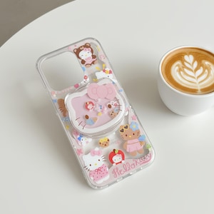 May include: Clear phone case featuring Hello Kitty and other cartoon characters. The case has a pink pop-up grip with a Hello Kitty face. The case is decorated with various pink, white, and brown cartoon figures. The words "Hello Kitty" are printed on the case.