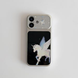 May include: Silver phone case with a black background featuring a white unicorn with wings. The top of the case is silver with a glittery finish and a small unicorn design. A stylish accessory.