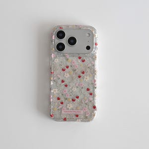 Könnte beinhalten: Transparente Handyhülle mit floralem Design. Die Hülle zeigt kleine rosa, weiße und gelbe Blumen sowie rote Erdbeerakzente. Die Hülle hat ein glitzerndes Aussehen und ein kleines rosa Etikett mit Text.