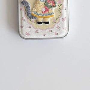 a small, white, floral-patterned case with a cat design on it, placed on a white surface.