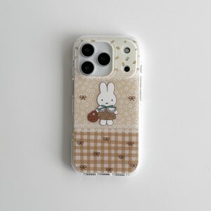 Könnte beinhalten: Transparente Handyhülle mit beigem Blumenmuster und einer Cartoon-Hasen-Illustration. Die Hülle hat unten ein braun-weißes Karomuster mit Schleifenakzenten. Der Hase trägt ein Kleid und hält eine braune Tasche.