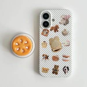 May include: A white phone case with a black polka dot pattern, adorned with various cartoon animal and food designs. A matching pop socket with a cookie design is also visible.