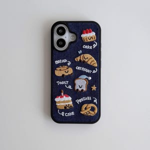 Może przedstawiać: Granatowe etui na telefon z haftowanymi wzorami jedzenia, w tym ciastem, rogalikiem, tostem i preclem. Etui ma czarną obwódkę i zawiera słowa "cake", "bread", "toast", "croissant" i "pretzel".