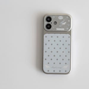 Könnte beinhalten: Silberne Handyhülle mit hellblauem Hintergrund und kleinen grauen Sternen. Der obere Bereich hat ein glitzerndes Silber-Finish mit Noten und dem Text "ピンクバンド". Der untere Bereich trägt den Text "Angel's wings".