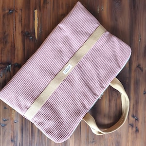 Peut inclure: Un sac fourre-tout en velours côtelé rose poussiéreux avec une sangle et des poignées beiges. Le sac a une forme rectangulaire et une fermeture éclair blanche. Le sac est sur une surface en bois.
