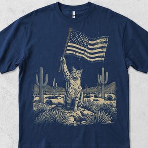 Puede incluir: Camiseta azul marino con un gato sosteniendo una bandera estadounidense en un paisaje desértico con cactus. El diseño desgastado tiene un aspecto vintage. La palabra "Navy" está impresa en la parte inferior de la camiseta.